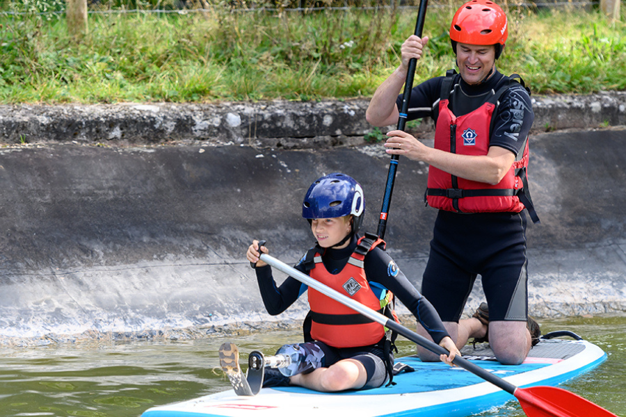 A young boy with a prosthetic leg sits on a paddle board with an adult, both rowing down a canal.